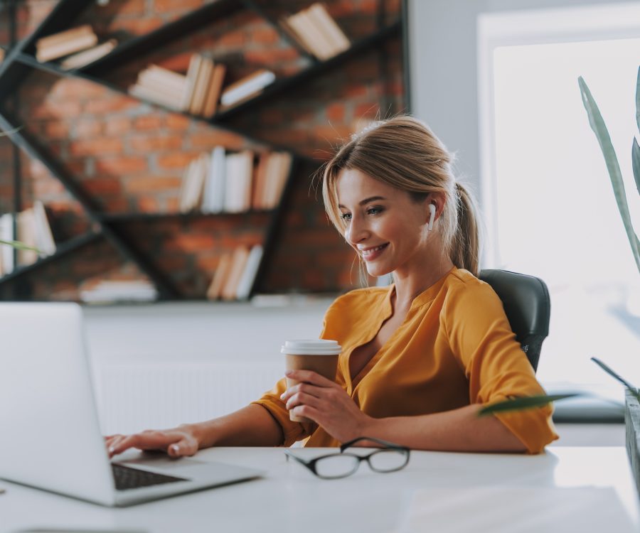 Blonde woman in an orange blouse having a carton cup of coffee in her hand and wearing wireless earphones while working on the laptop